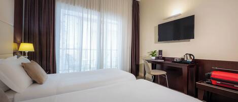 A hotel room with two beds, a desk with a chair, a TV mounted on the wall, and a window with curtains.