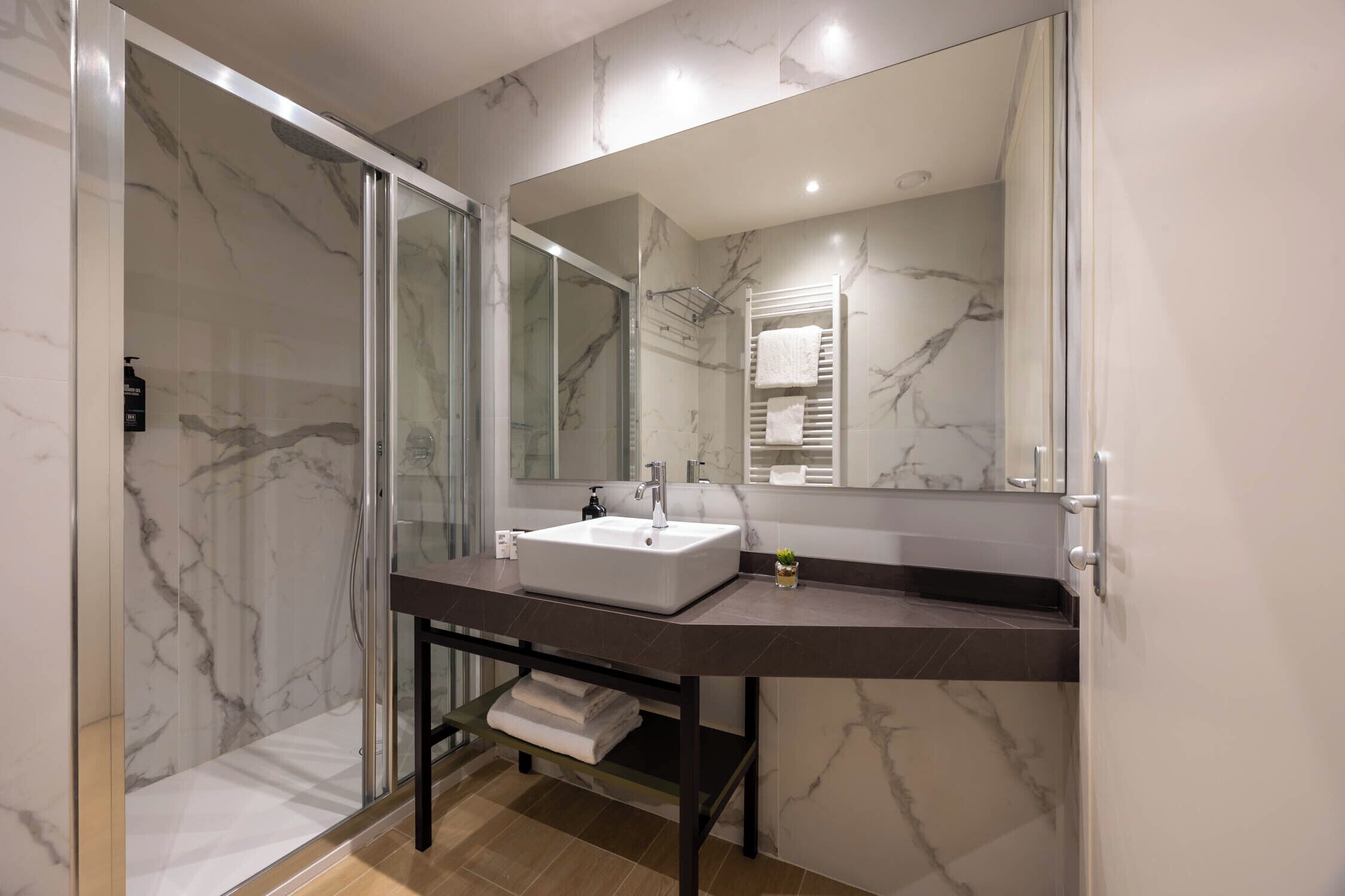 A modern bathroom with a glass-enclosed shower, a marble-tiled wall, a white sink, and a large mirror.