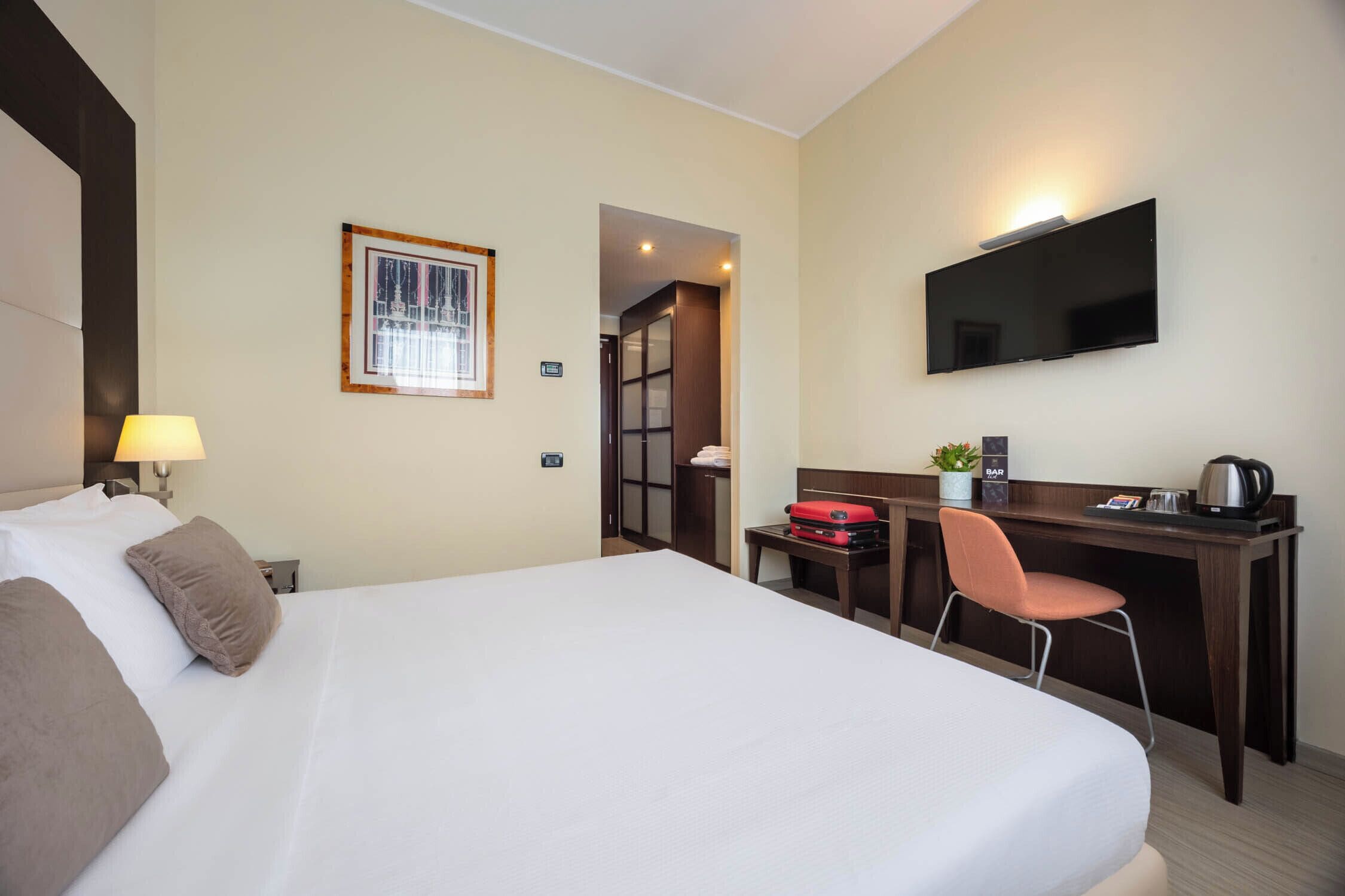 Habitación de hotel con una cama grande, un escritorio con silla, un televisor y una mesita con una planta y una tetera.