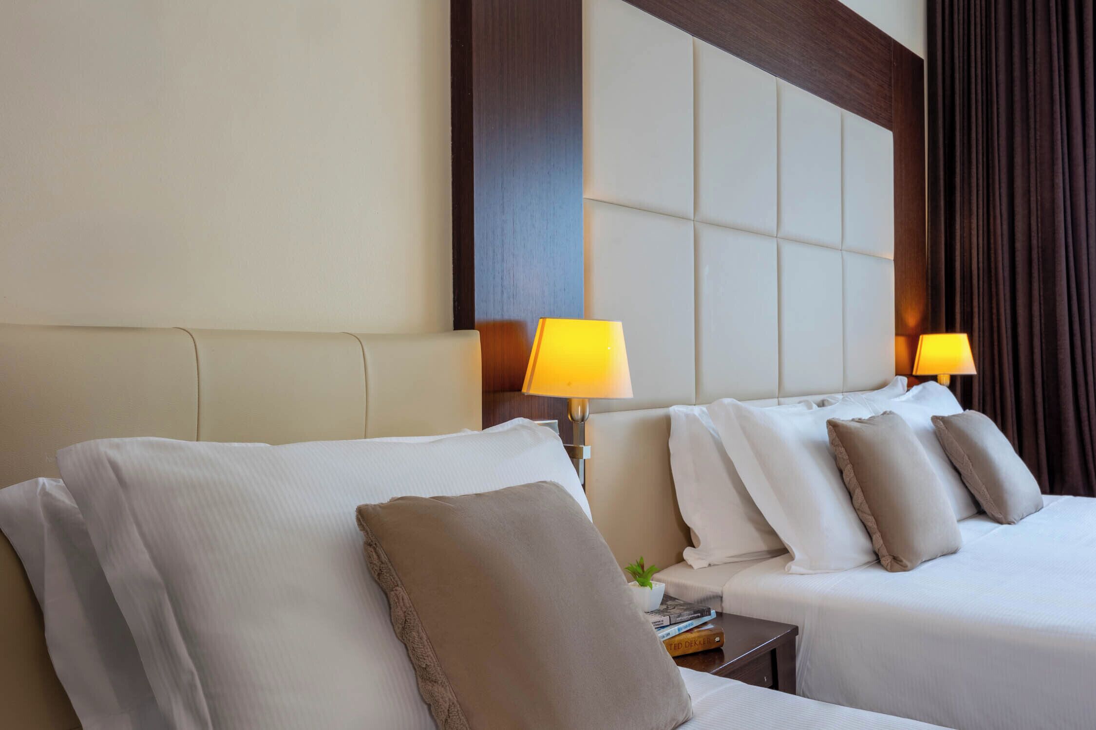 Une chambre d’hôtel avec deux lits, chacun agrémenté de draps blancs et de plusieurs oreillers. On y trouve deux lampes de chevet avec des abat-jour jaunes. La chambre est dotée d’une grande tête de lit rembourrée et d’un mur d’accent en bois foncé.