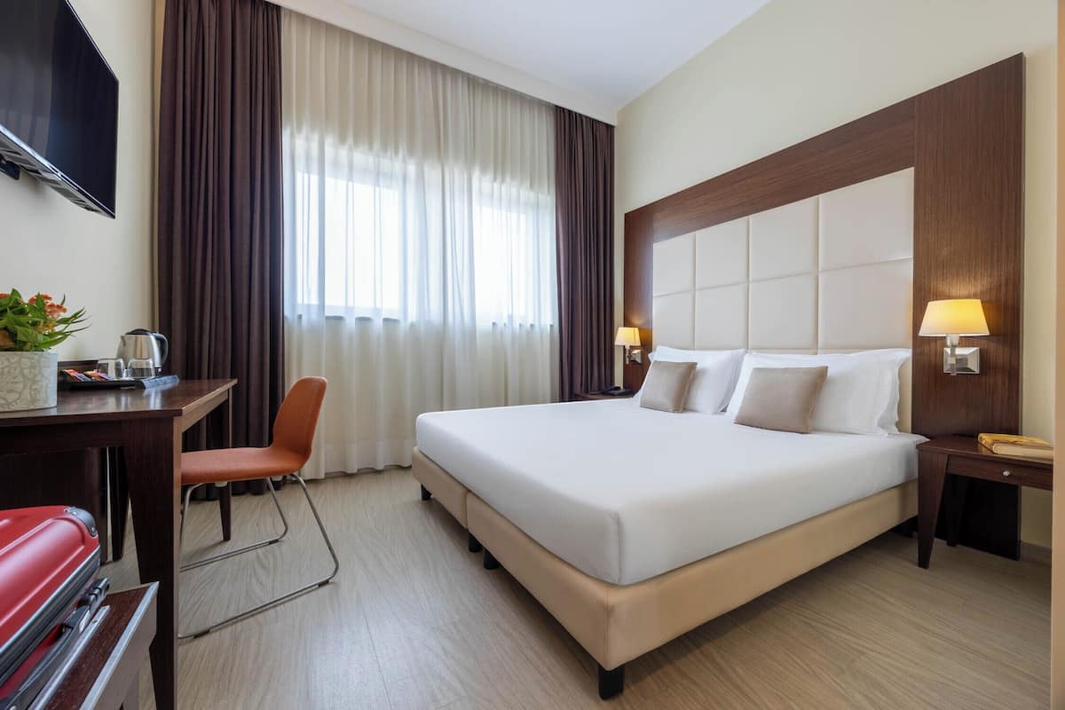 Ein Hotelzimmer mit einem großen Bett, einem Schreibtisch mit Stuhl, einem an der Wand befestigten Fernseher und einem Fenster mit Vorhängen.