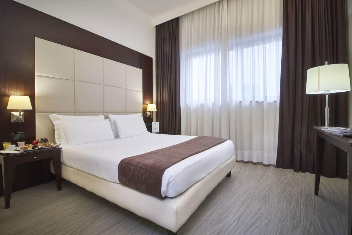 Ein Hotelzimmer mit einem großen Bett, zwei Nachttischen mit Lampen, einem Fenster mit Vorhängen und Blick nach draußen.