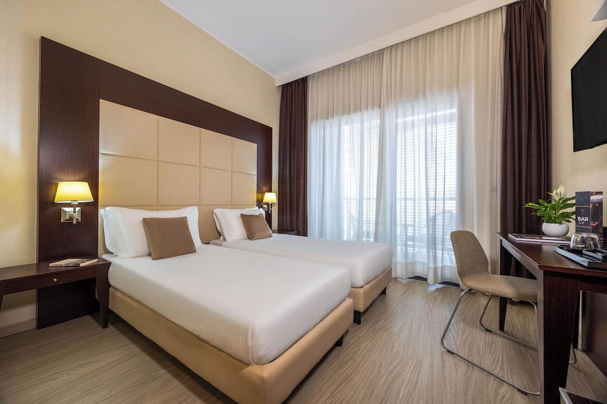 Ein Hotelzimmer mit zwei Betten, einem Schreibtisch, einem Stuhl, einem Fernseher und einem Fenster mit Vorhängen.