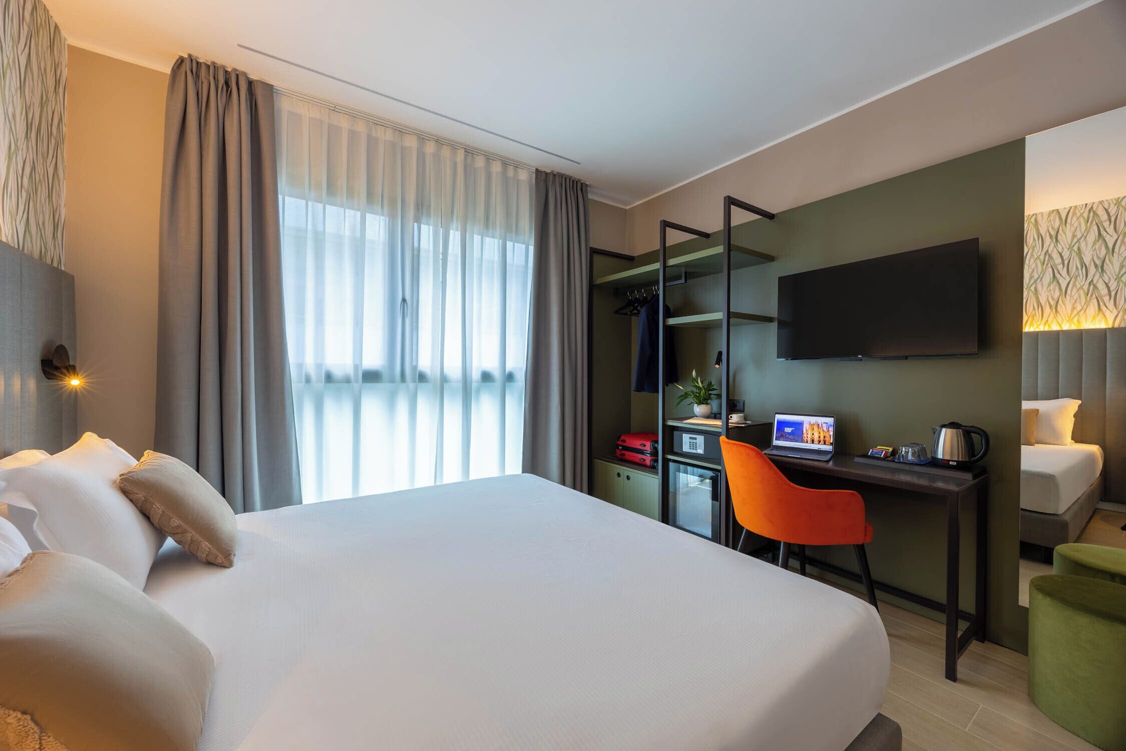 A hotel room with a large bed, a desk with a laptop, a TV mounted on the wall, and a window with curtains.