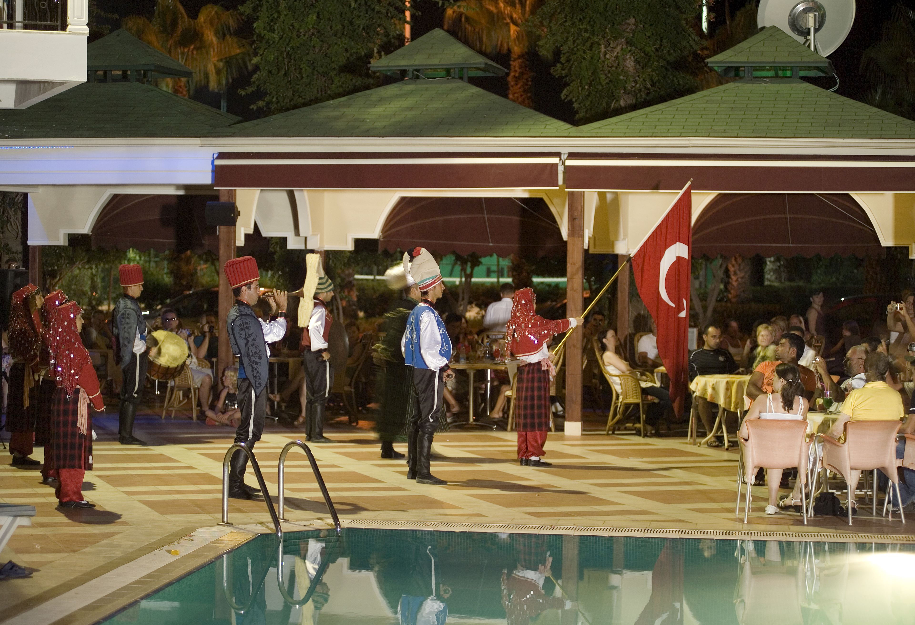 A poolside area with a checkered floor, outdoor seating, and a stage with performers.