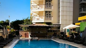 Una piscina con sillones de playa y sombrillas, rodeada por un edificio de varios pisos con balcones.