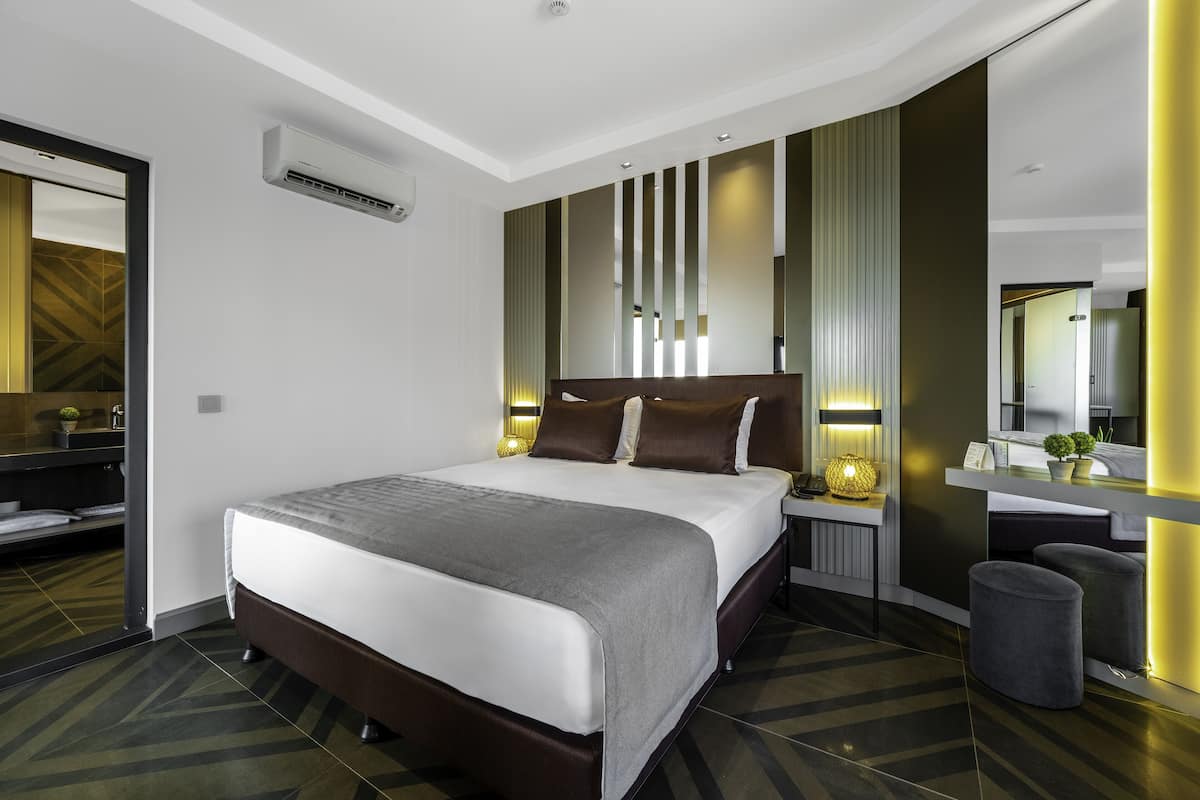 A modern hotel room with a large bed, a bedside table with a lamp, a mirror, and a view of the bathroom.