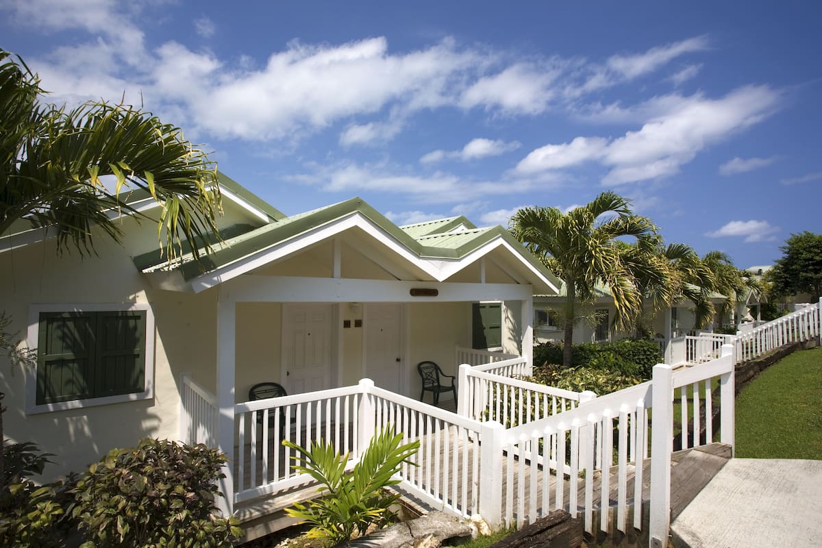 A row of white houses with green roofs and white railings, surrounded by palm trees and a clear blue sky.