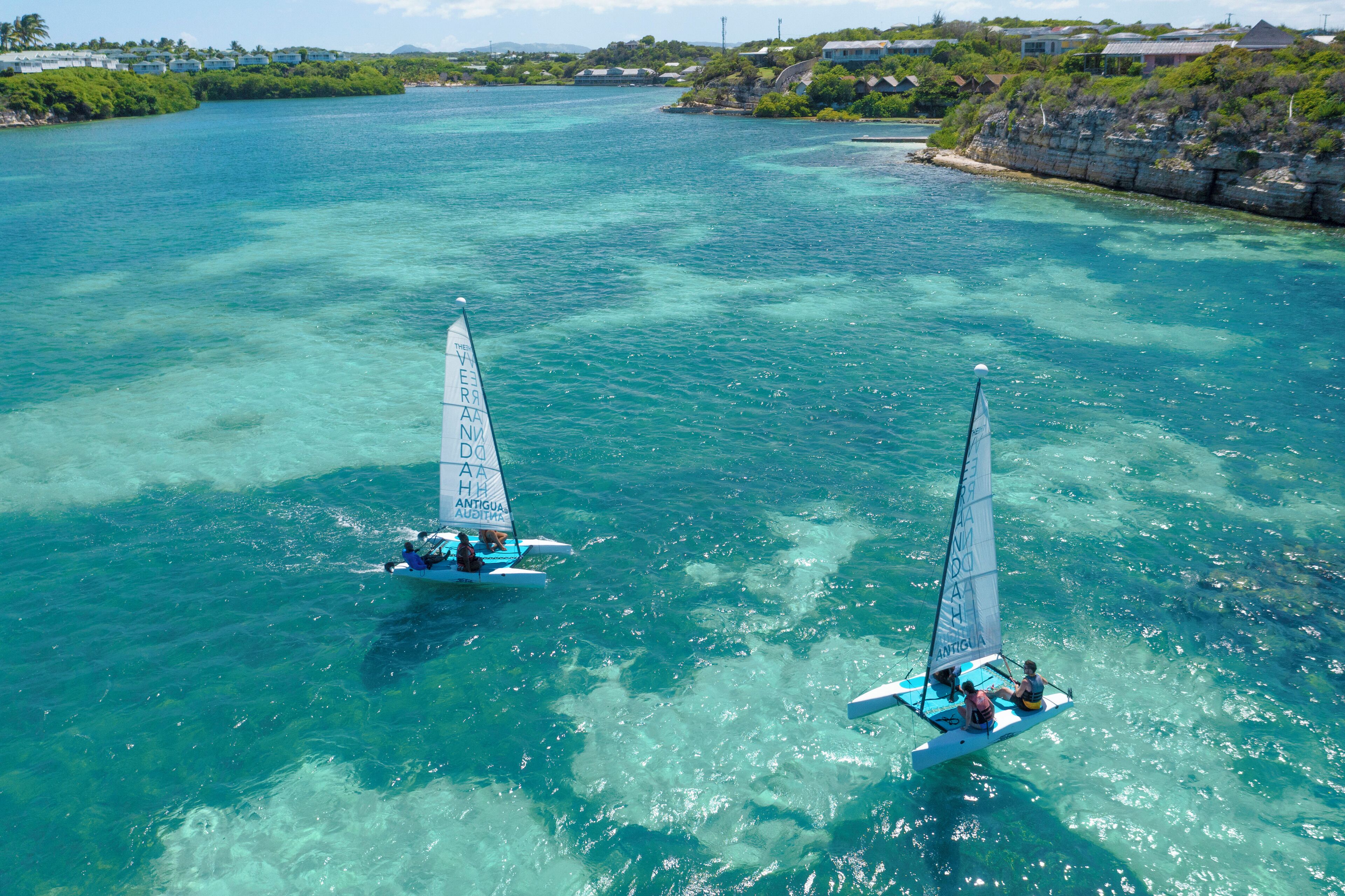 Two sailboats on a clear turquoise sea with a rocky coastline and buildings in the background.