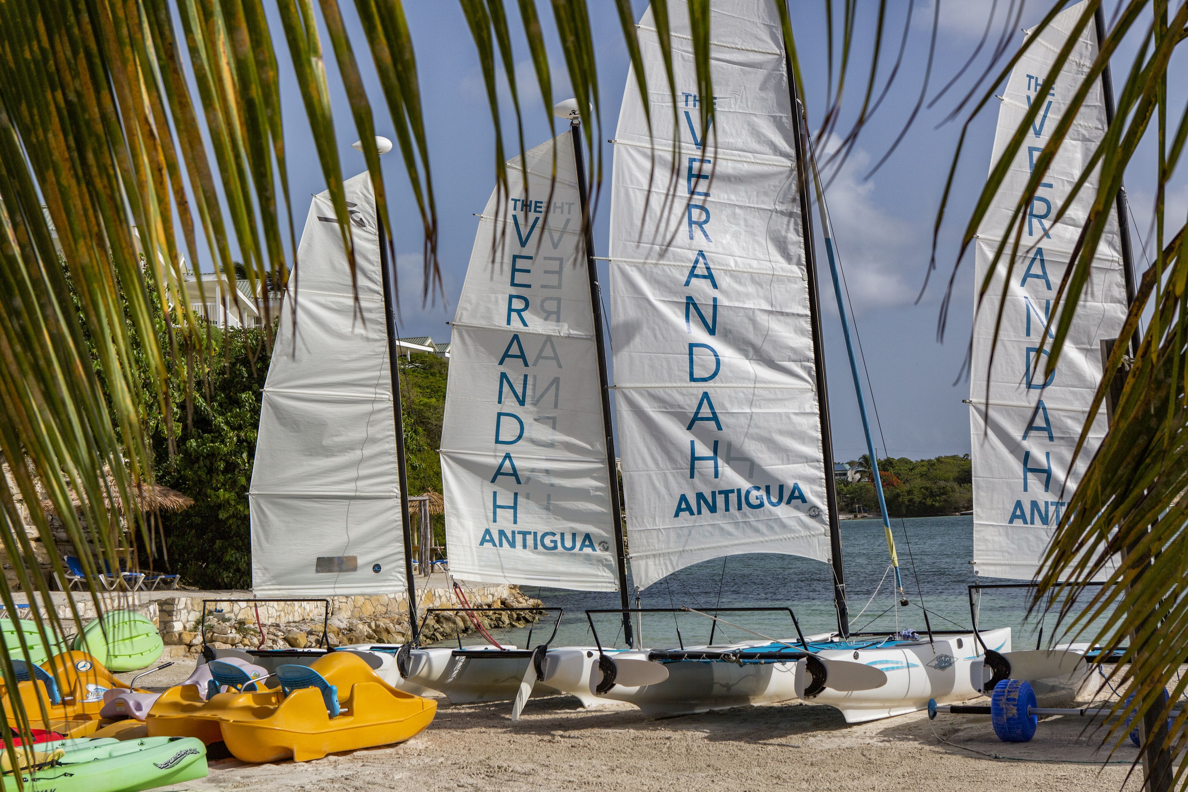 Una playa con varios veleros amarrados, algunos con velas que dicen 'VERANDAH ANTIGUA'.