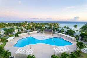 A large, curved swimming pool with lounge chairs and umbrellas, surrounded by palm trees and a view of the ocean.