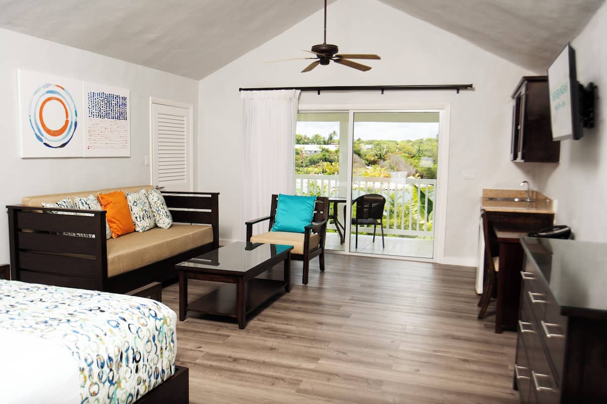 A modern room with a bed, a sofa, a coffee table, a chair, a desk, and a television. There is a sliding glass door leading to a balcony with a view of greenery.