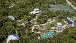 Vista aérea de un complejo turístico con piscina, canchas de tenis y diversas zonas ajardinadas.