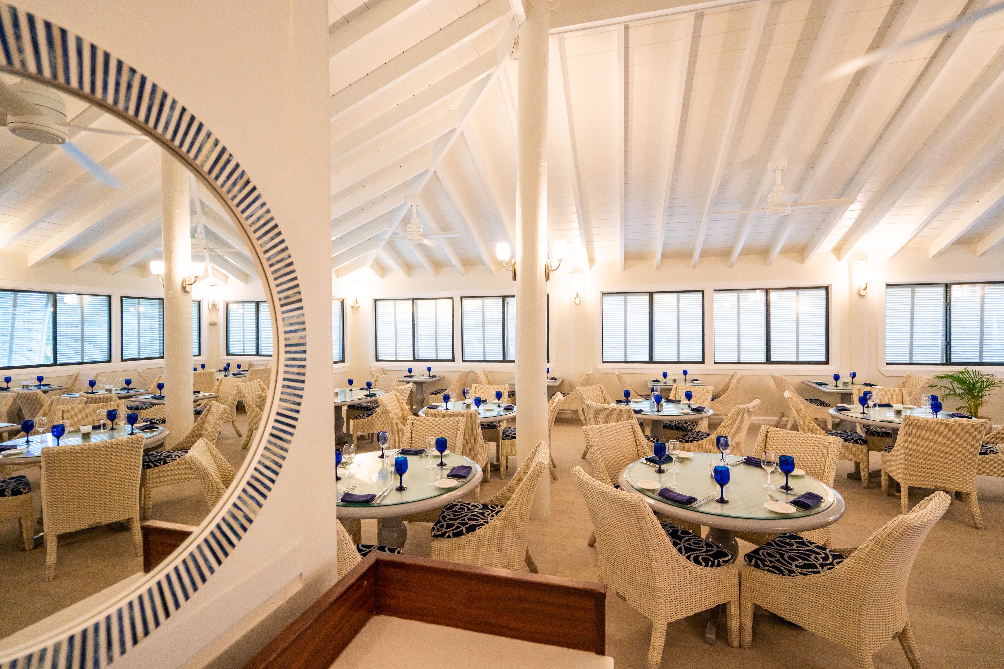 Un espace repas avec des tables rondes et des chaises en osier, de grandes fenêtres et un plafond haut avec des poutres apparentes.