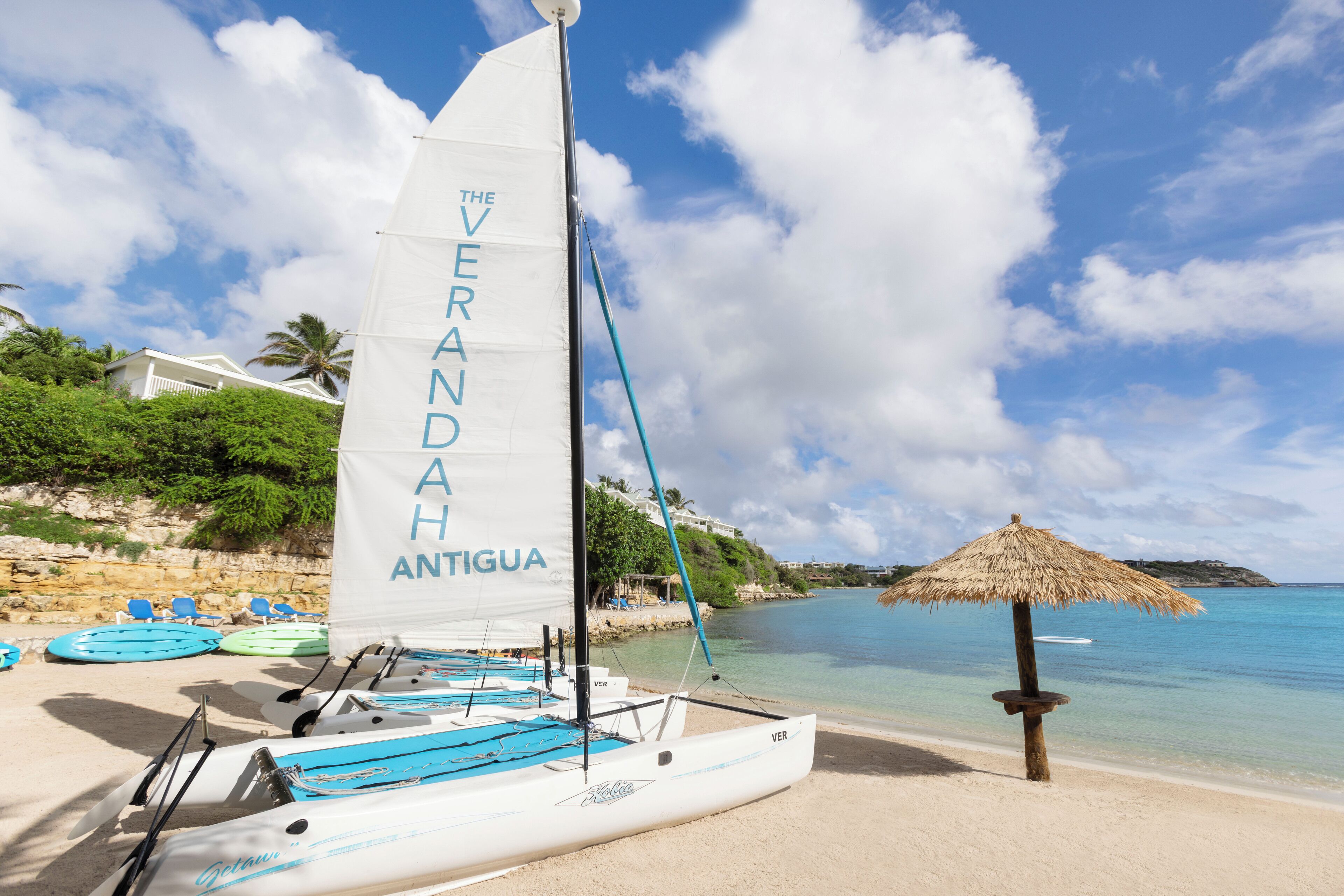 A sailboat named "The Veranda Antigua" on a sandy beach with clear blue water and a thatched umbrella nearby.