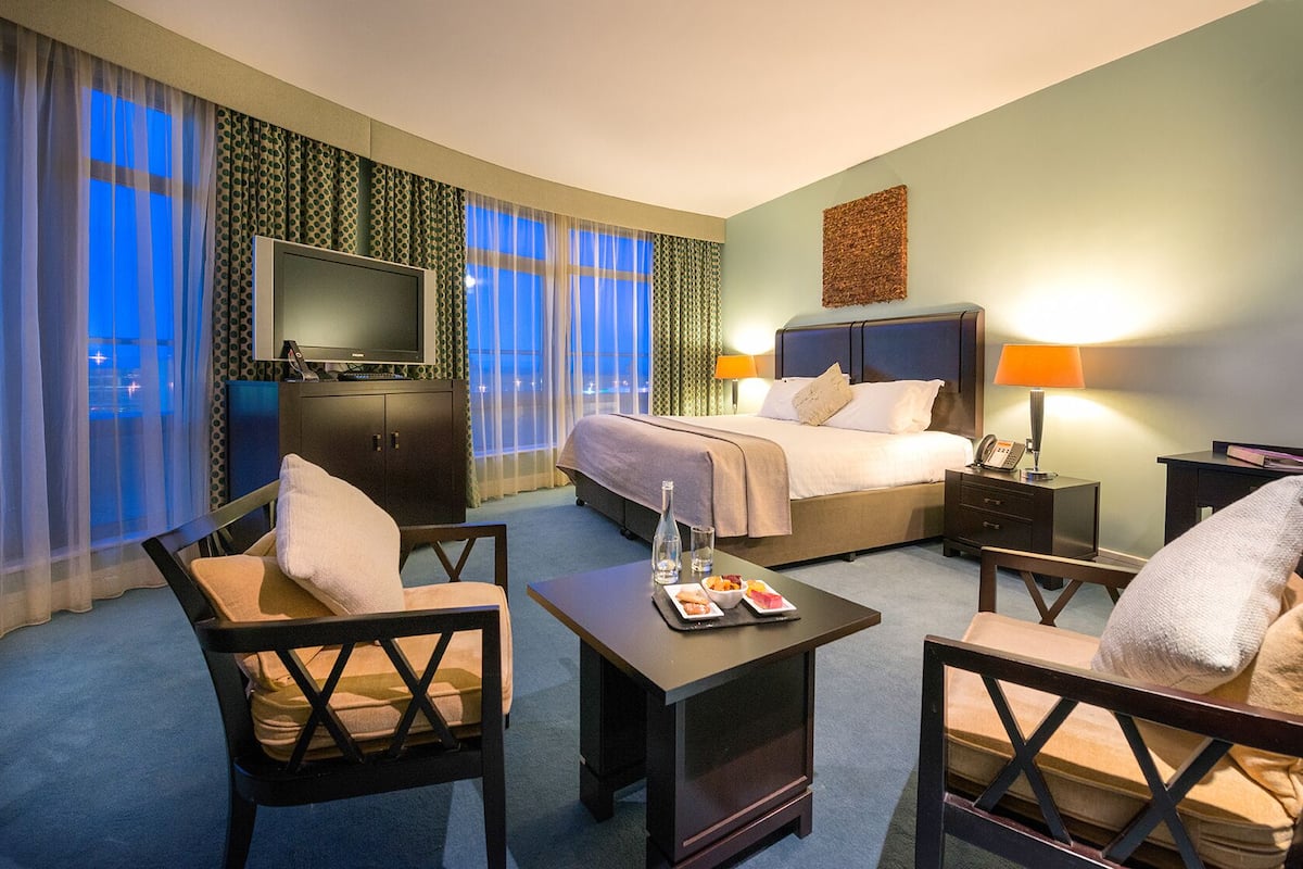 A hotel room with a bed, two chairs, a desk with a lamp, a TV, and a minibar.