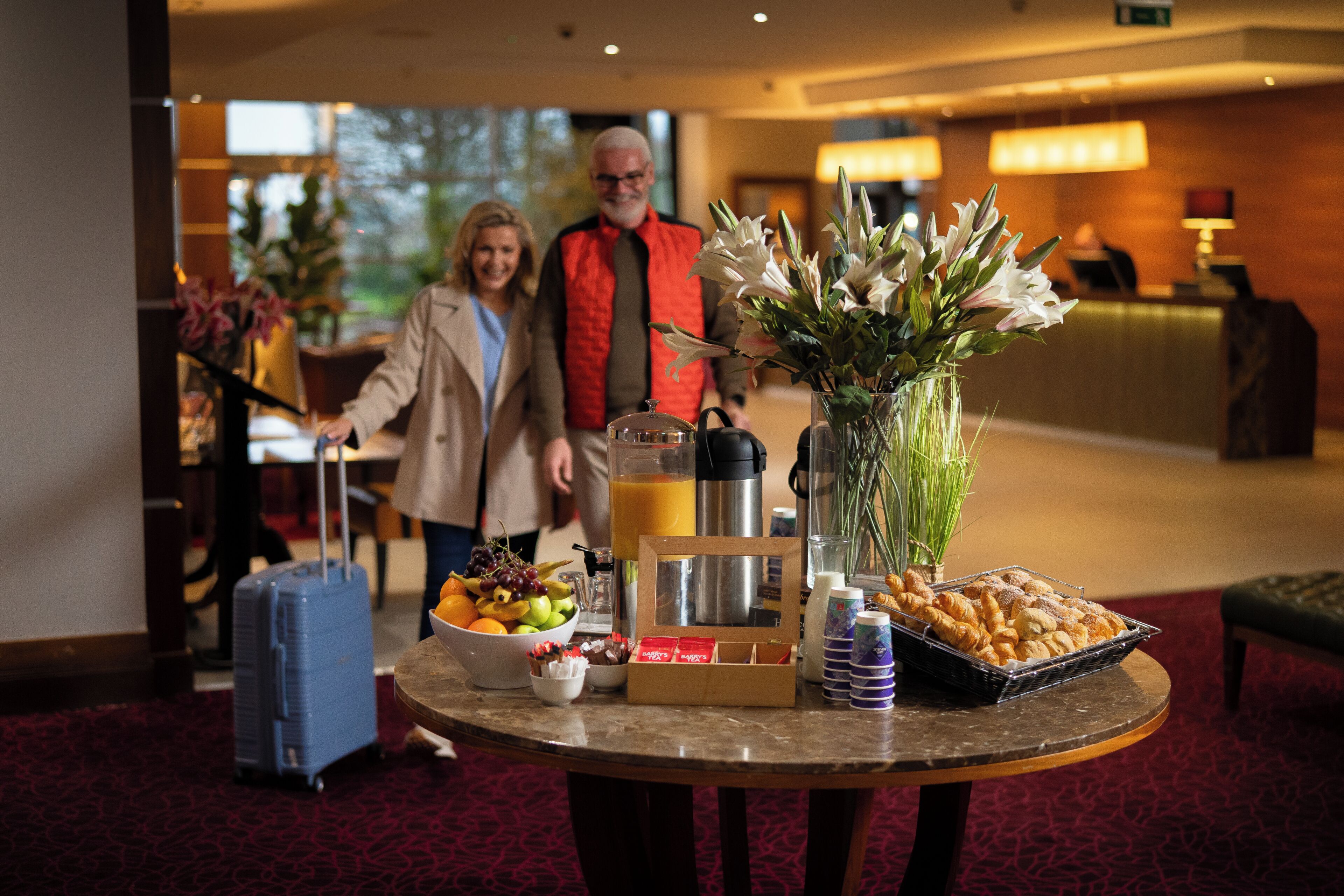 Un vestíbulo de hotel con un mostrador de recepción, una mesa con alimentos para el desayuno y una maleta azul.