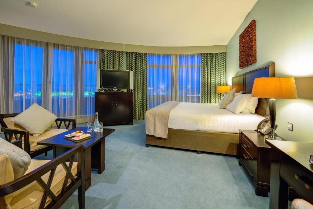 Quarto de hotel com cama, escrivaninha, cadeira, televisão e vista para a cidade à noite.