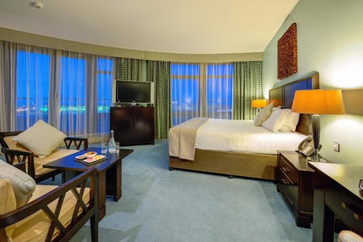 Quarto de hotel com cama, escrivaninha, cadeira, televisão e vista para a cidade à noite.