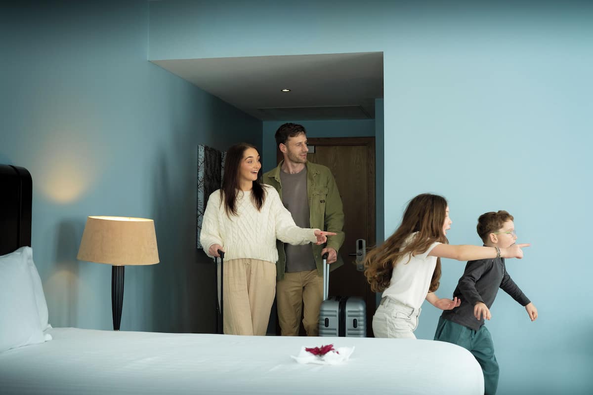 A hotel room with a bed, a lamp, a suitcase, and a door.