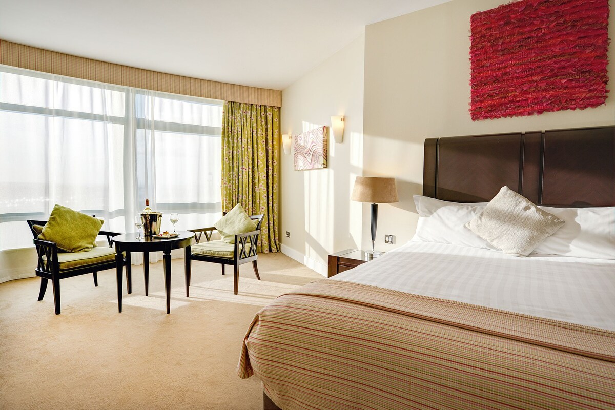 A hotel room with a large bed, a dining table with chairs, a view of the sea, and a red abstract artwork on the wall.