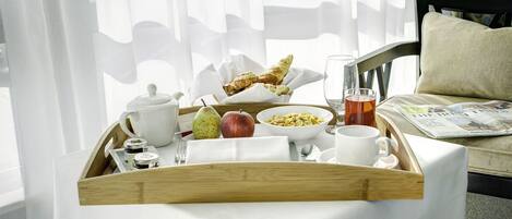Ett frukostbricka med en päron, ett äpple, en skål med flingor, en kopp te och en tidning på ett bord nära ett fönster med tunna gardiner.