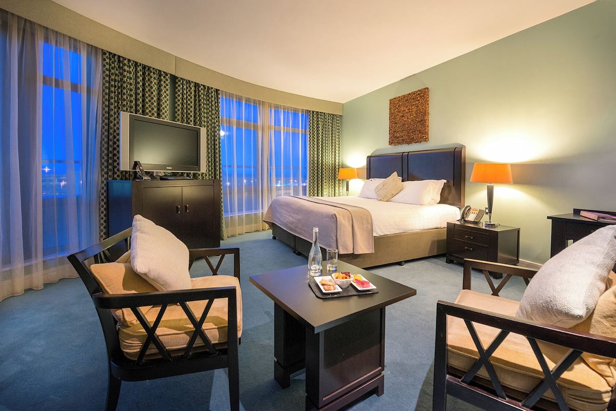 A hotel room with a bed, two chairs, a desk with a lamp, a TV, and a minibar.