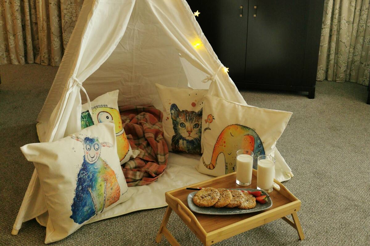 A cozy indoor setting with a white tent adorned with fairy lights, a wooden tray with cookies and milk, and colorful pillows.