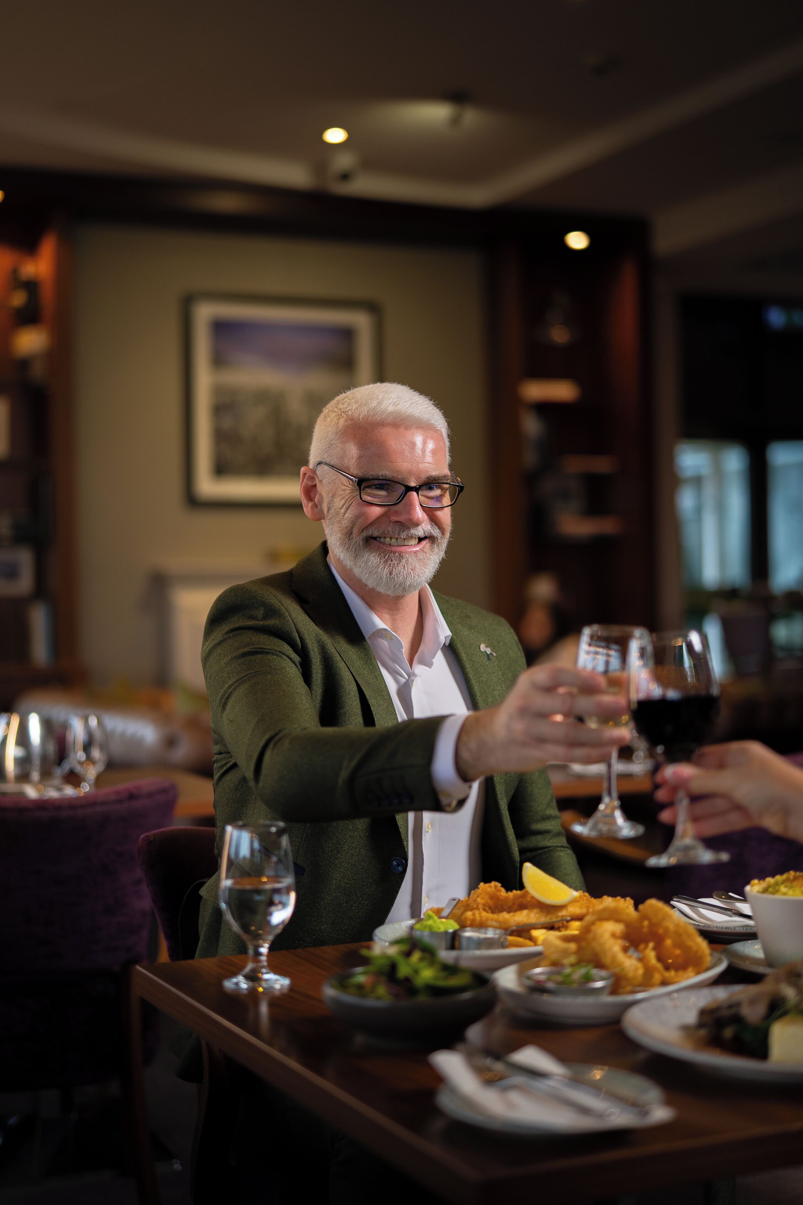 A man seated at a table with a glass of red wine, a bowl of food, and a glass of water.