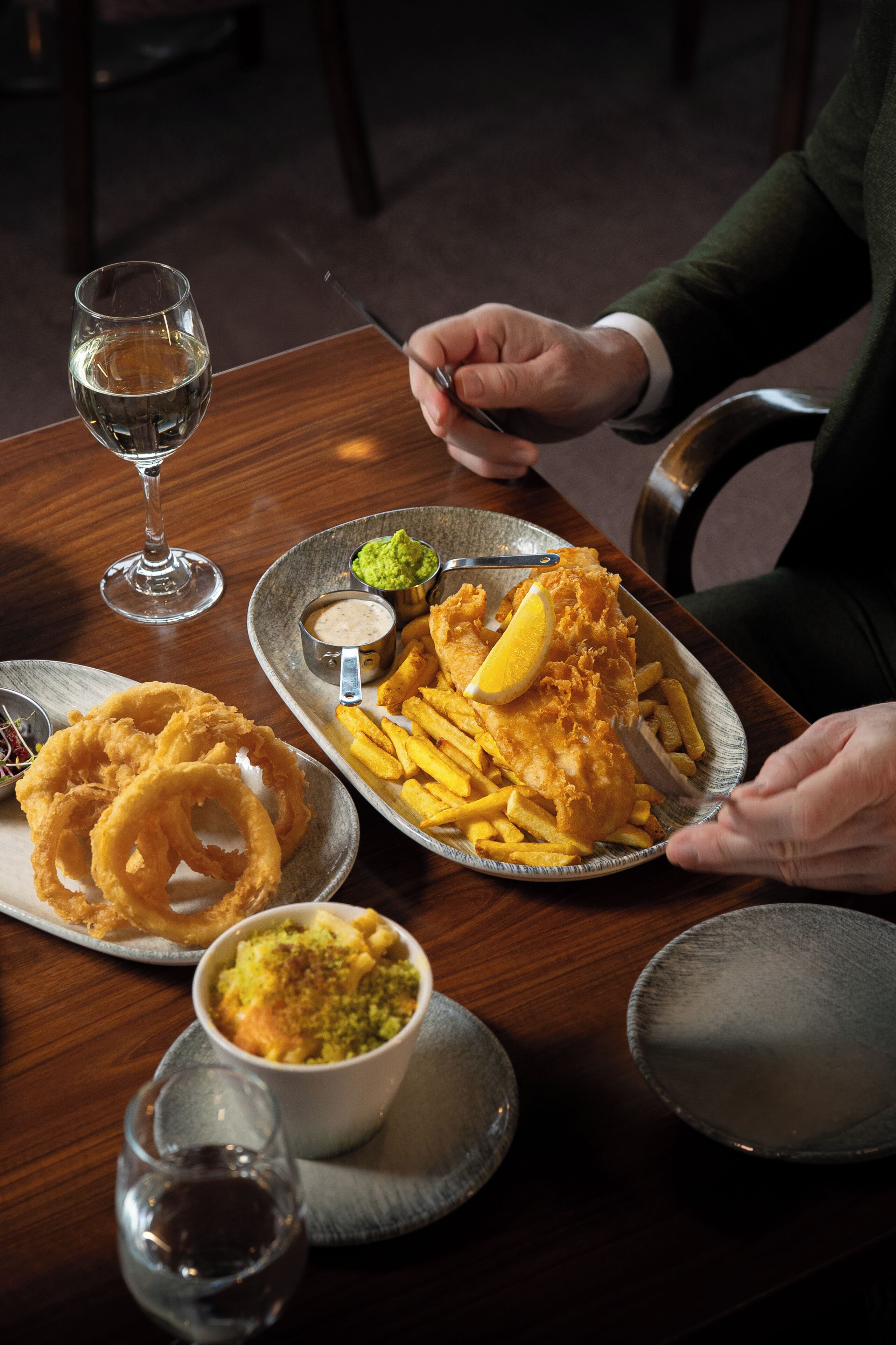 A dining table with a plate of fish and chips, onion rings, and a small bowl of sauce.