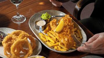 A dining table with a plate of fish and chips, onion rings, and a small bowl of sauce.