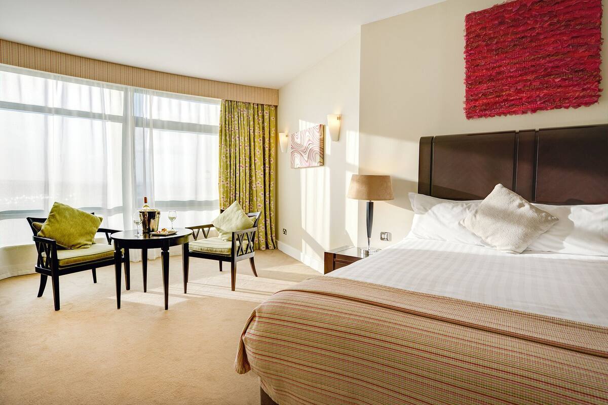 Quarto de hotel com uma cama grande, uma mesa de jantar com cadeiras, uma mesinha com abajur e vista para o mar por grandes janelas.