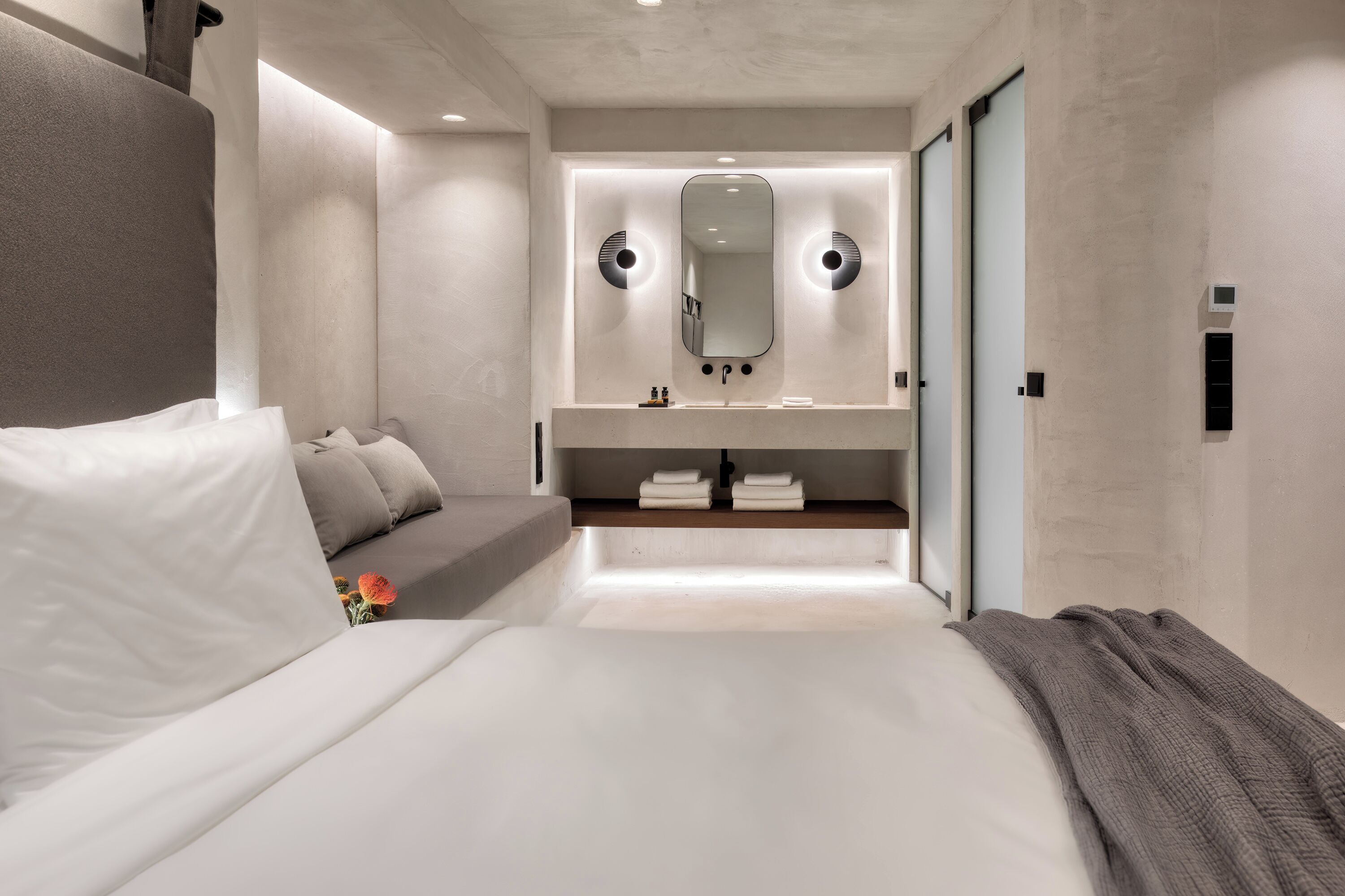 A modern hotel room with a large bed, a bedside table, a mirror, a built-in shelf with towels, and a sliding door.