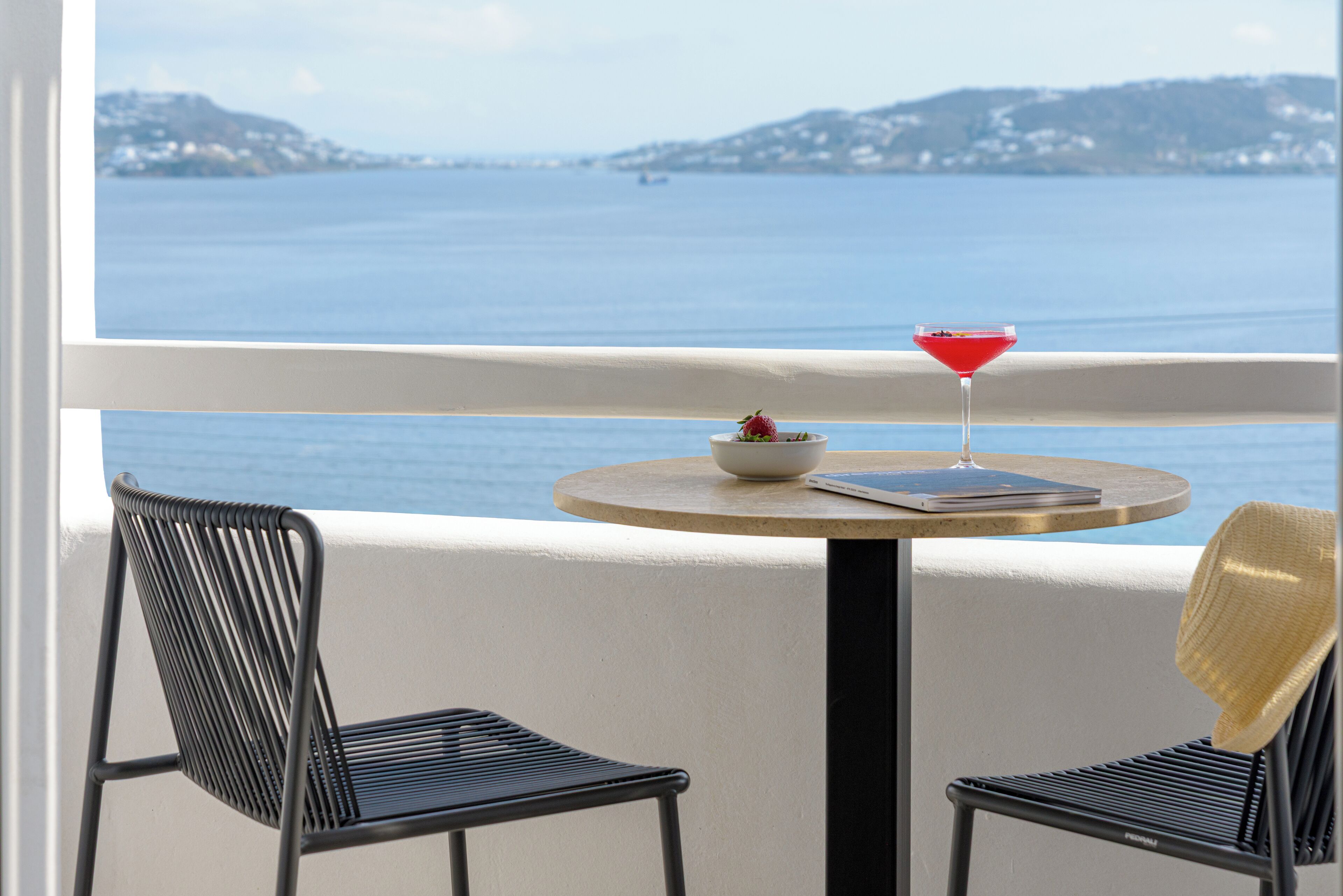 A balcony with a table, two chairs, a red drink, a bowl with fruit, and a view of the sea and distant hills.