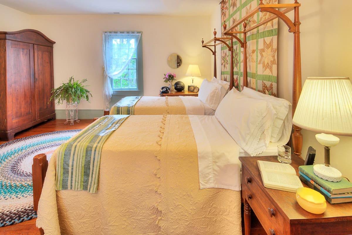 A bedroom with a large bed, a wooden wardrobe, a bedside table with books and a lamp, and a window with curtains.