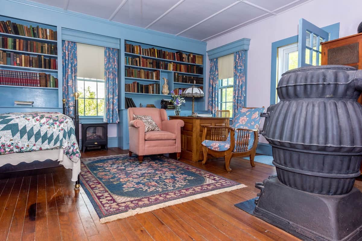 A room with wooden flooring, a bed with a floral bedspread, a pink armchair, a wooden desk, a large black stove, and bookshelves filled with books.