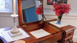A wooden vanity with a mirror, a white lamp, and a vase with pink flowers. - Oak Grove Bed and Breakfast (South Boston)
