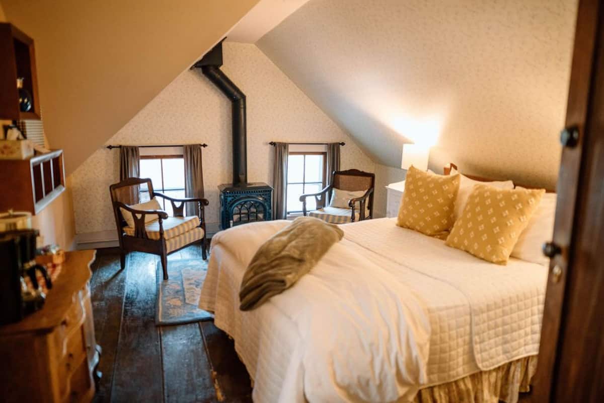 Un dormitorio acogedor en el ático, con techo inclinado, cama con ropa de cama blanca, dos sillas, una mesita y una chimenea.