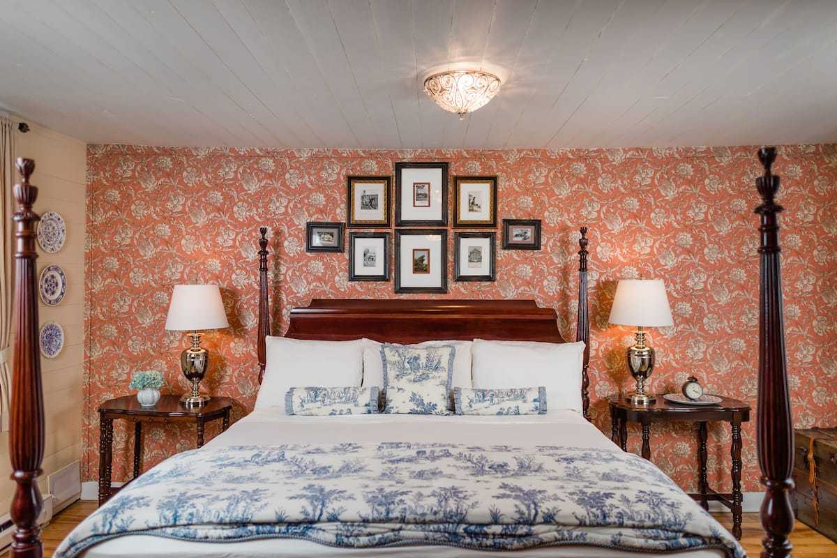 Un dormitorio con una cama de madera con dosel, dos mesitas de noche con lámparas, un papel tapiz floral y cuadros enmarcados en la pared.