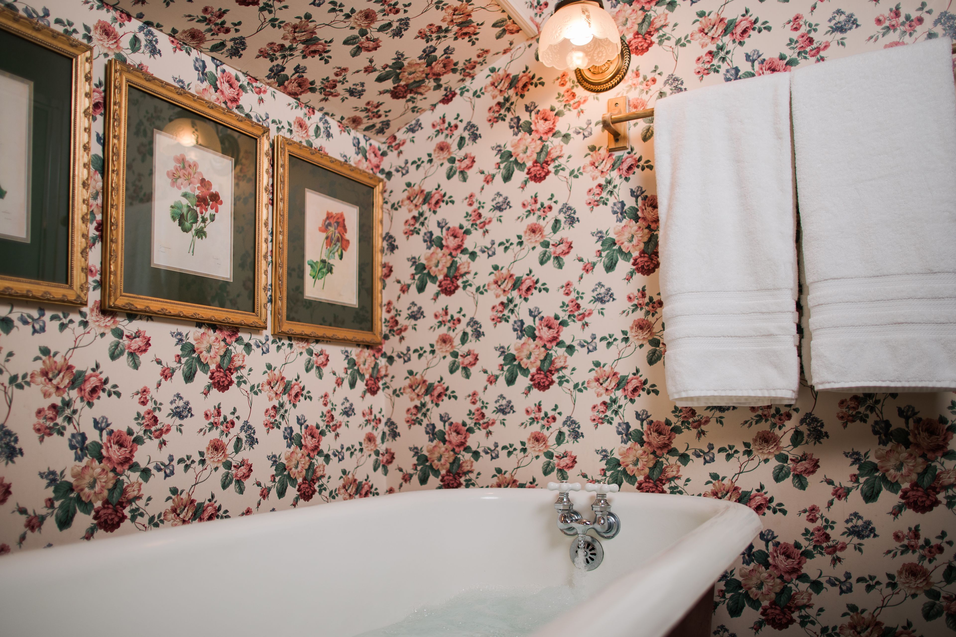 A bathroom with floral wallpaper, a bathtub, framed floral pictures, and a towel rack.