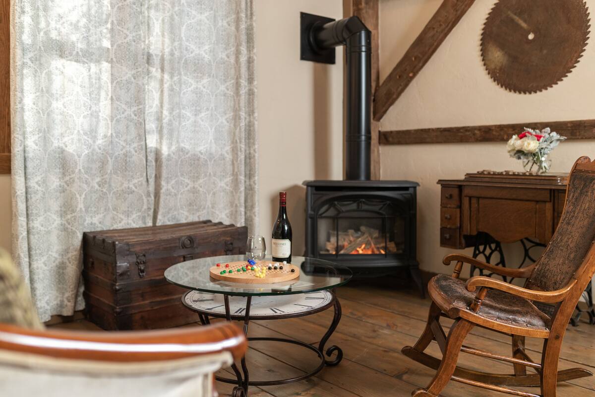 Una habitación acogedora con un cofre de madera, una mesa de vidrio con un reloj, un sillón mecedora, una chimenea y un jarrón con flores.