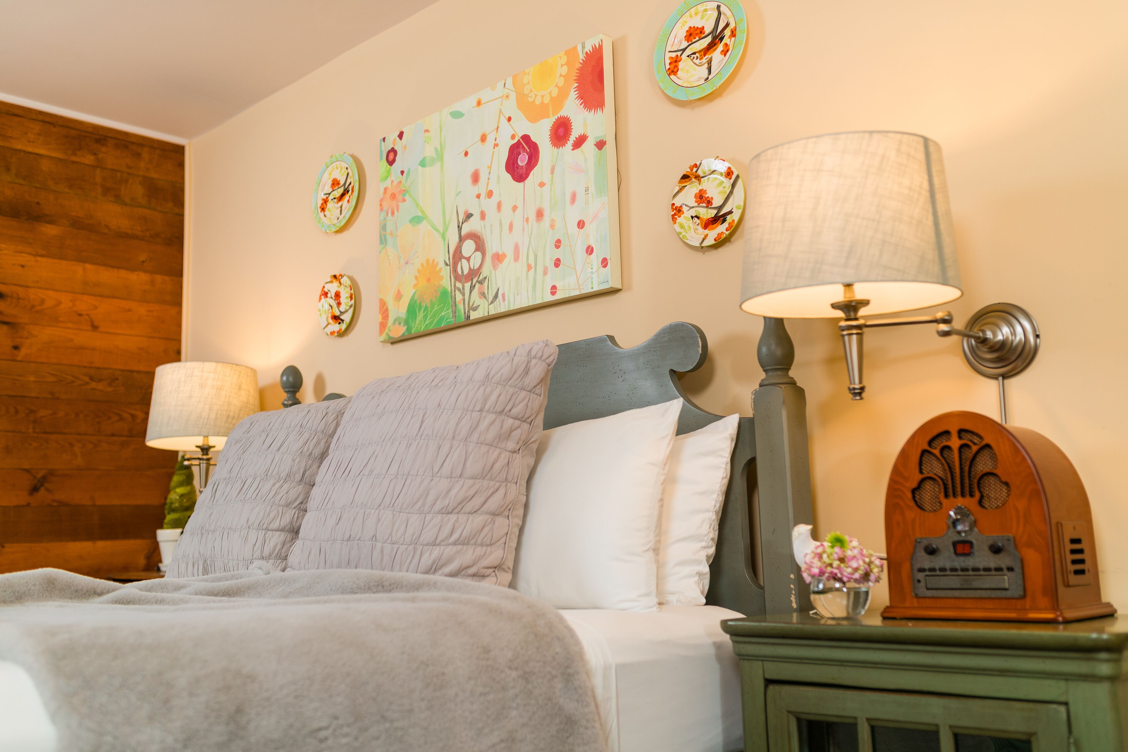 Une chambre à coucher avec un lit, deux lampes fixées au mur, une radio et un tableau représentant des fleurs.