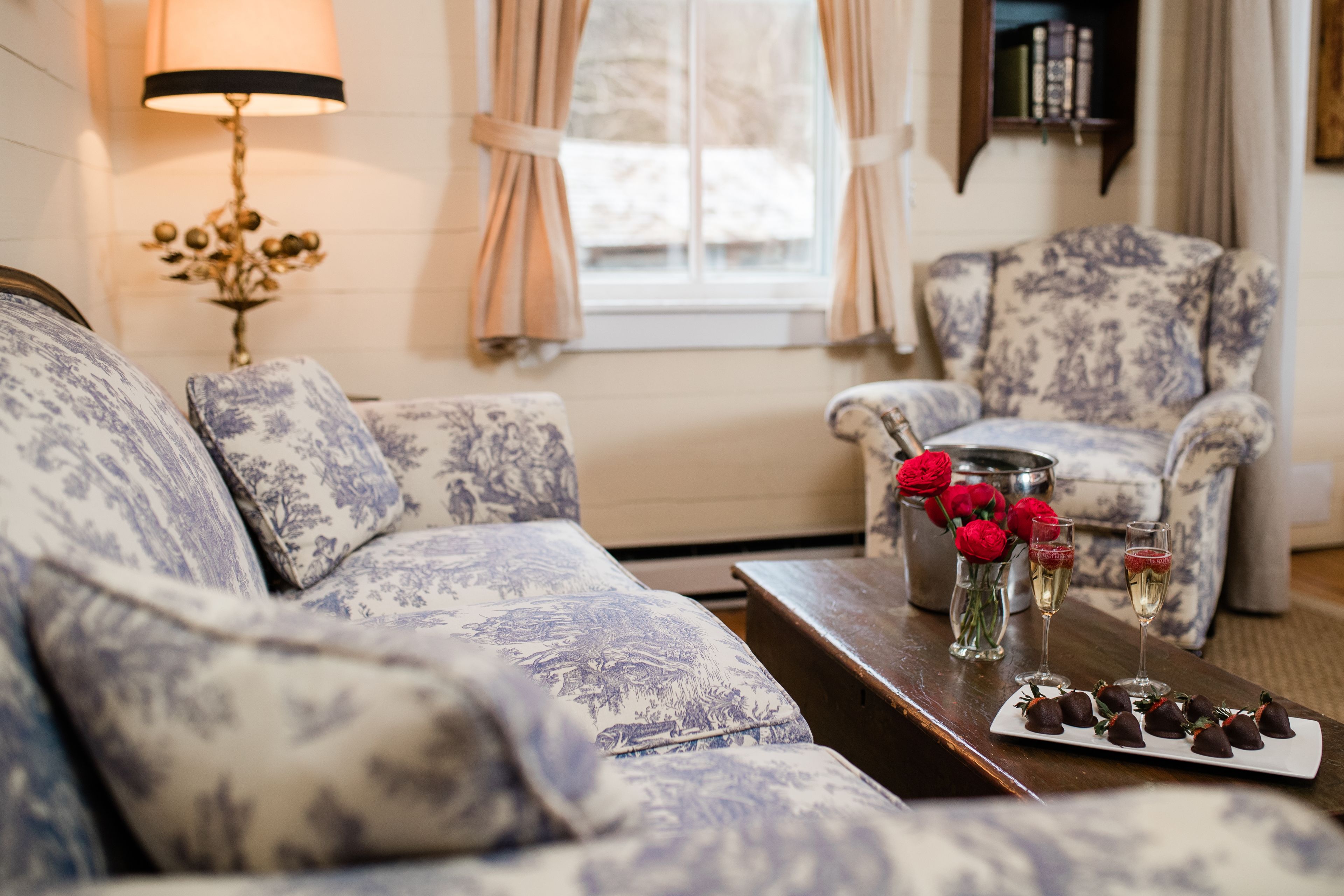 A living room with patterned sofas, a coffee table with chocolates and drinks, and a vase with red roses.