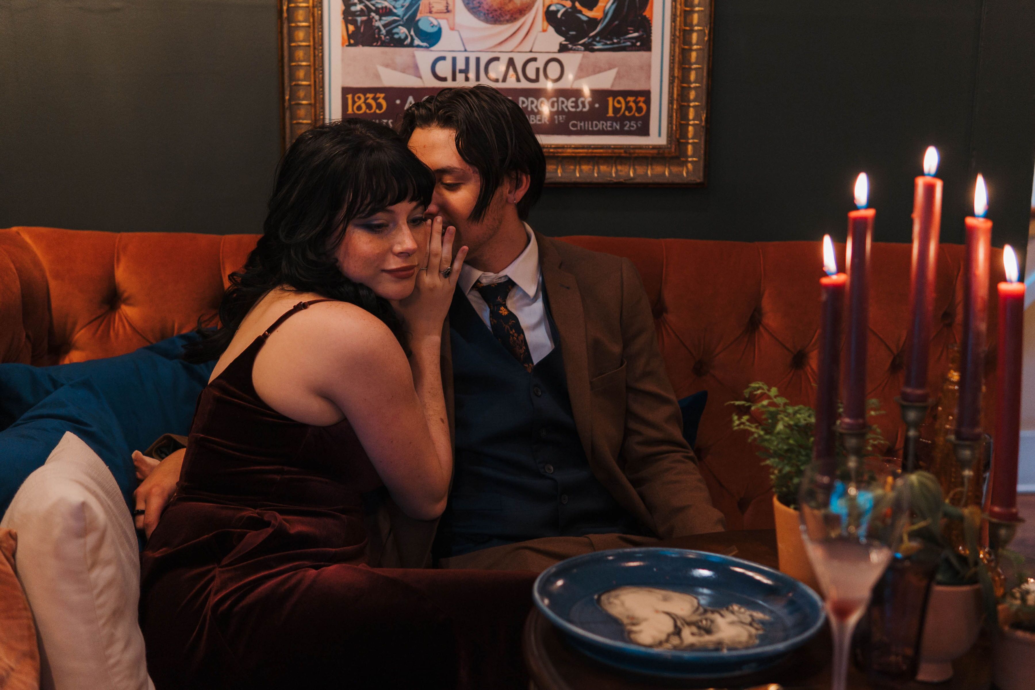 A couple sitting on a tufted sofa in a warmly lit room with a vintage poster on the wall and a candelabra with lit candles on a table.