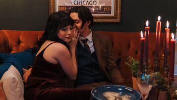 A couple sitting on a tufted sofa in a warmly lit room with a vintage poster on the wall and a candelabra with lit candles on a table.