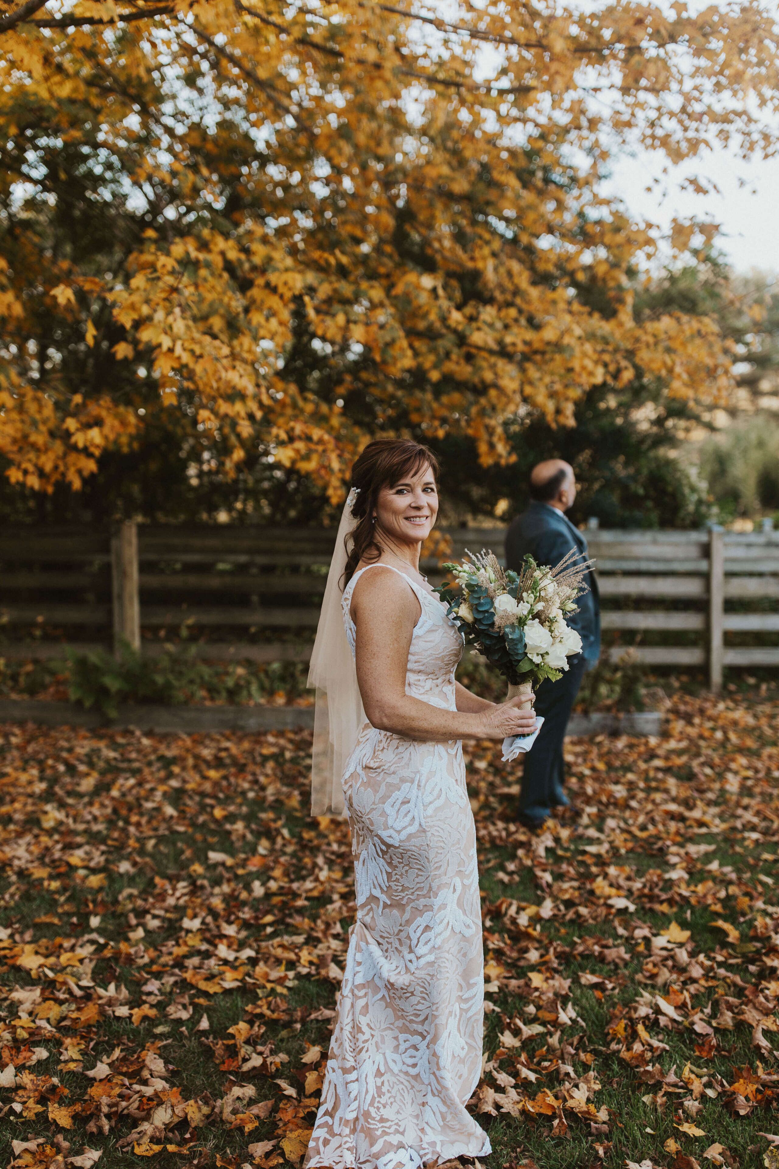 A bride in a lace dress holding a bouquet, standing on a leaf-covered ground with a wooden fence and trees in the background.