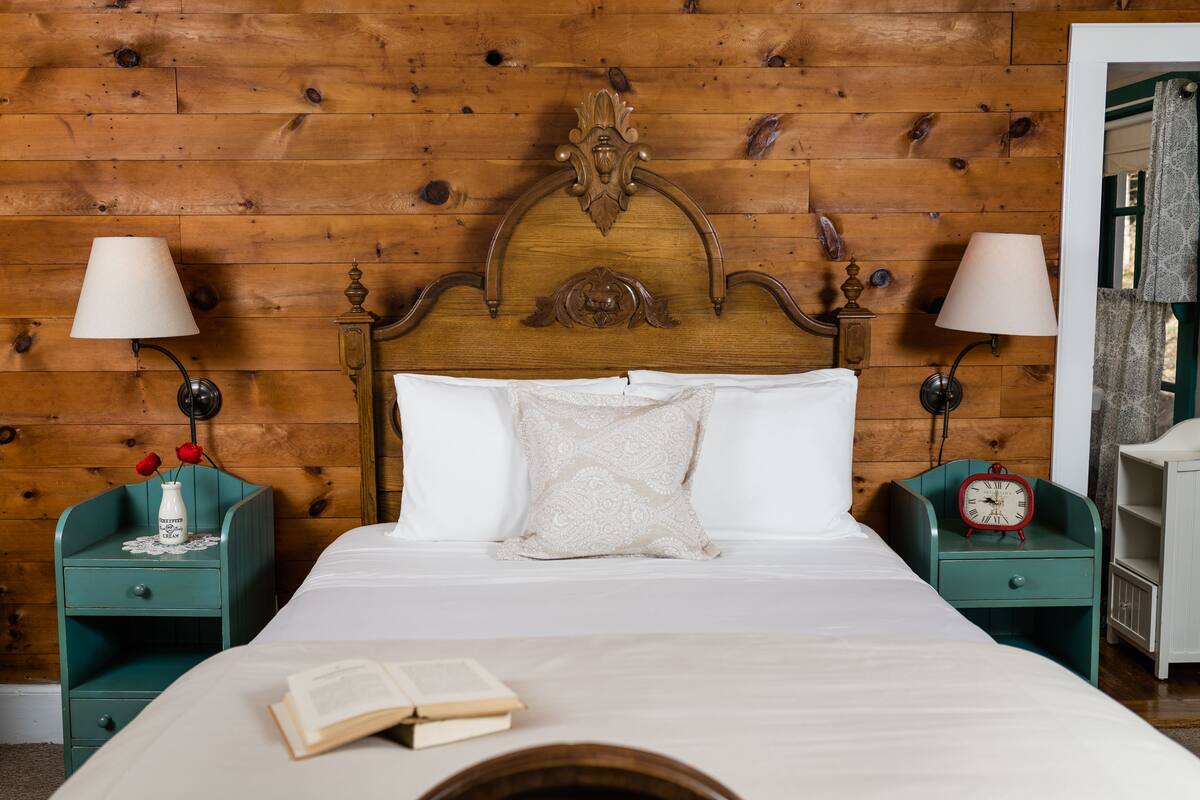 Habitación con paneles de madera, una cama, dos mesitas de noche, un reloj rojo y un libro sobre la cama.