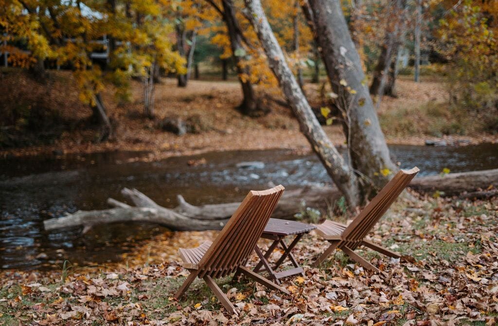 Deux chaises en bois, placées au bord d’une rivière, avec des feuilles d’automne jonchant le sol.