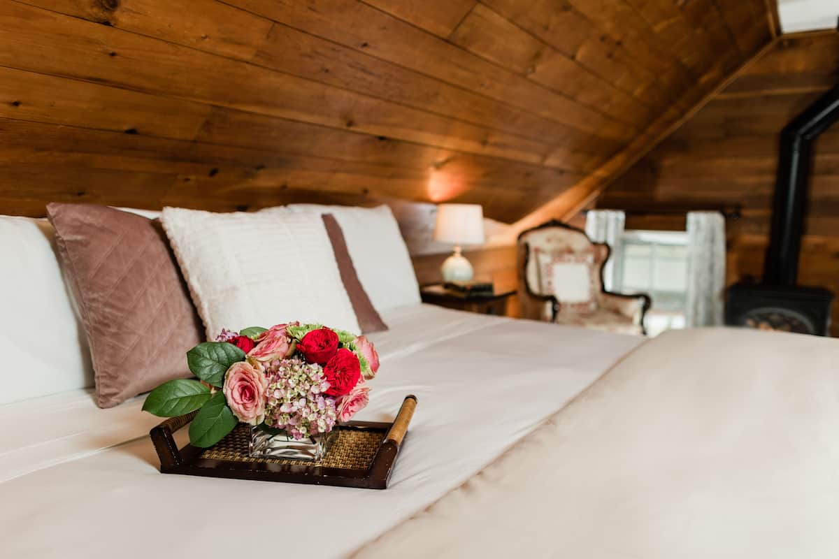 Un dormitorio de cabaña de madera con cama, almohadas, una lámpara de noche y una bandeja con flores.