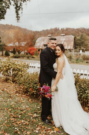 A couple in wedding attire standing in a garden with a white fence and autumn leaves on the ground.